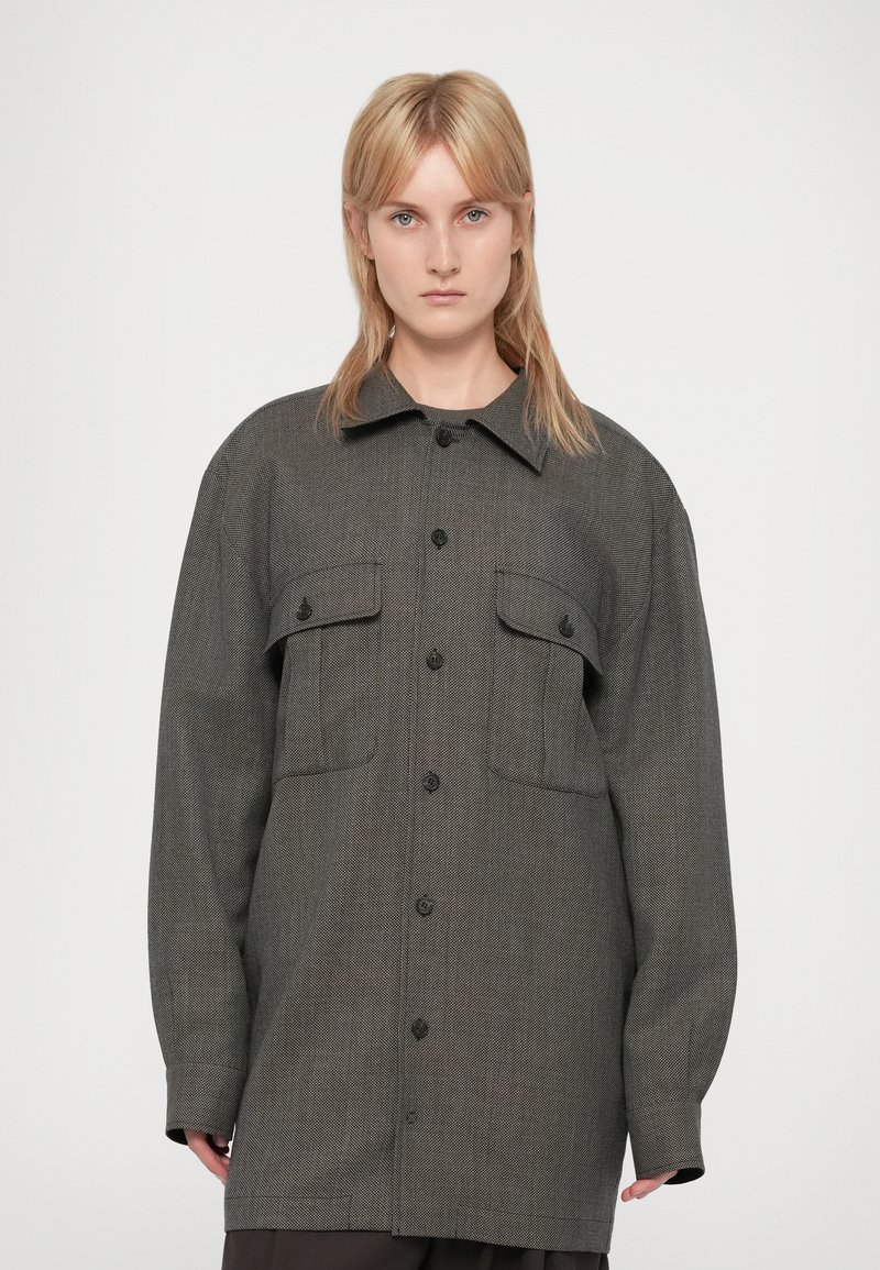 Mujer con cabello rubio lacio que lleva una chaqueta gris de botones oversized con dos bolsillos en el pecho, contra un fondo blanco liso.