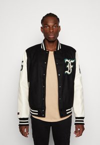 Black and white varsity jacket with a front F logo, striped cuffs and waistband, snap button closure, and soft texture. Worn over a beige shirt.