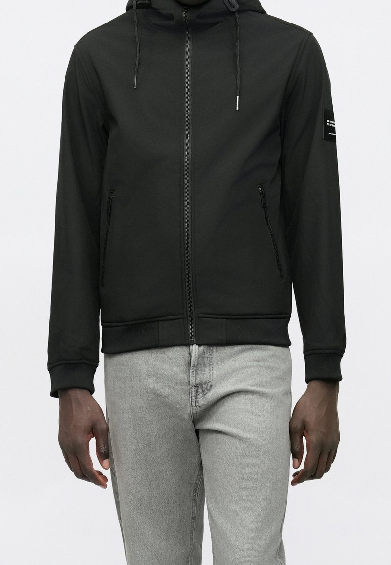 Sweat à capuche noir zippé avec cordons et poches latérales zippées, porté avec un jean gris clair, montré sur une personne du cou jusqu’aux hanches.