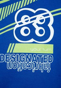 T-shirt in cotone blu con grafica testuale bianca e verde, che include "FABBRICAZIONE DESIGNATA" e elementi di design circolari.