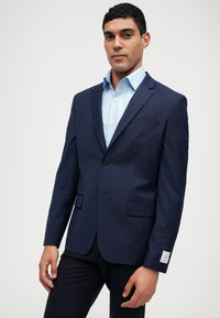 Tamsiai mėlynas kostiumo švarkas iš glotnaus audinio su vienu klinkiu, notch tipo apykakle ir dviem priekinėmis kišenėmis. Po juo šviesiai mėlynas marškinys.