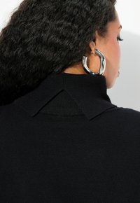Maglione nero a maglia con colletto alto e design con schiena aperta, caratterizzato da una texture liscia. Grande orecchino a cerchio in argento visibile di profilo.