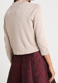 Beige cropped sweater with a high neckline, three-quarter sleeves, and button accents. Worn over a textured burgundy skirt.
