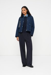 Casaco acolchoado azul-marinho com fecho éclair e botões de pressão, apresentando um bolso no peito. Combinado com umas calças largas escuras e um suéter de malha texturizada.