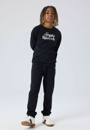 Jeune personne avec des dreadlocks portant un sweat-shirt noir "Empty Spaces", un pantalon noir et des baskets blanches, debout devant un fond clair uni.