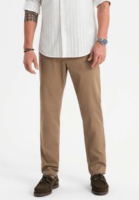 Pantalons en coton beige à coupe droite, avec une texture subtile, portés avec des chaussures en daim marron foncé et une chemise à boutons à rayures claires.