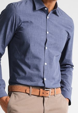 Homme portant une chemise bleue à manches longues boutonnée, rentrée dans un pantalon beige avec une ceinture marron texturée, mains dans les poches.