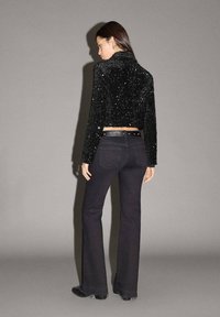 Giacca nera cropped con paillettes texturizzate, abbinata a jeans neri svasati. L'outfit presenta una silhouette aderente e una cintura a contrasto.