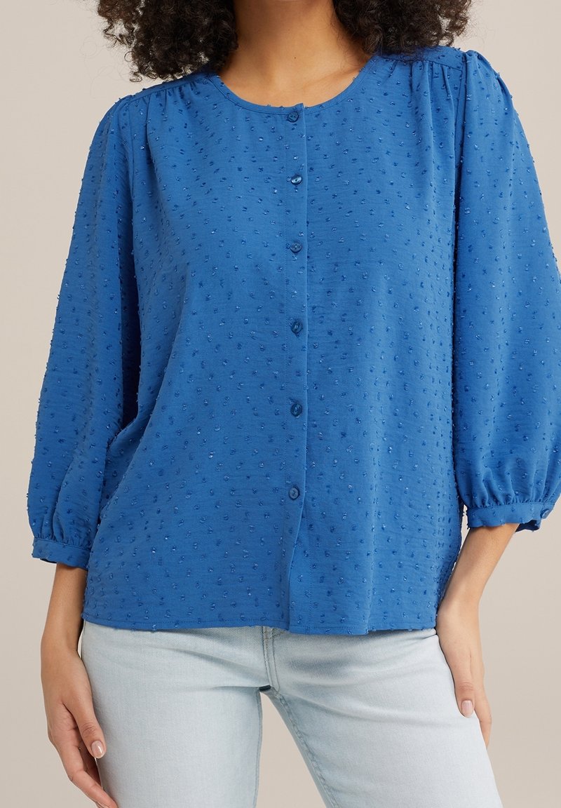 Femme portant un chemisier bleu texturé à boutons avec des manches bouffantes trois-quarts et un jean bleu clair, sur fond neutre.