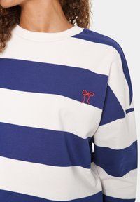 Gestreiftes Langarmshirt in Blau und Weiß aus Baumwolle, mit einem roten bestickten Schleifendetail im linken Brustbereich.