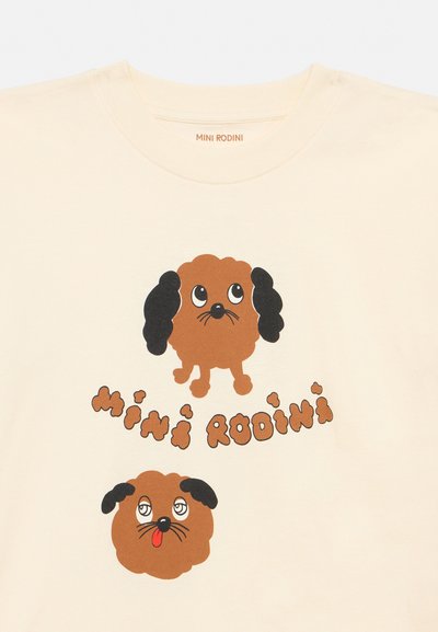Camisa color crema con dos caras de perros marrones de dibujos animados, una con los ojos mirando hacia arriba, la otra con la lengua fuera, y el texto "Mini Rodini" entre ellas.
