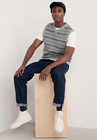 Grijze gebreide vest met blauwe en bruine patronen, gedragen over een witte T-shirt. Gecombineerd met donker denim jeans en witte high-top sneakers.