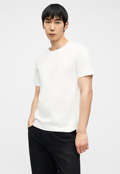 Νέος άνδρας με ίσια μαύρα μαλλιά, φοράει λευκό κοντομάνικο μπλουζάκι και μαύρο παντελόνι, στέκεται με το ένα χέρι στην τσέπη.