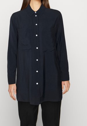 Chemise noire à manches longues avec boutons blancs, portée sur un pantalon noir, montrée des épaules jusqu'à mi-cuisse sur une personne debout.