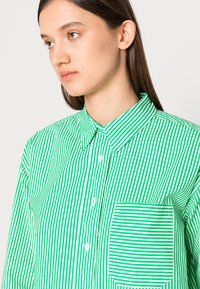 Camisa de botão às riscas verdes e brancas com colarinho, apresentando um bolso do lado esquerdo, feita de tecido leve.