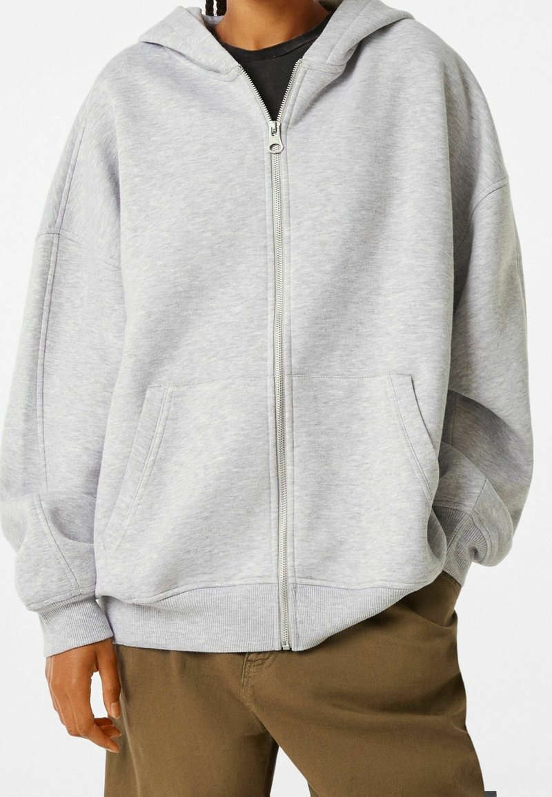 Persona vistiendo una sudadera con cremallera gris claro sobre una camiseta negra, combinada con pantalones marrones, con la mano en el bolsillo, recortada en el cuello y los muslos.
