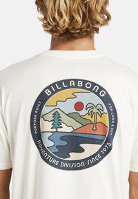 Weißes T-Shirt mit einem kreisförmigen grafischen Motiv von Bergen, Bäumen, einer Sonne und einem Fluss in Pastellfarben; der Text lautet "BILLABONG" und "ADVENTURE DIVISION SEIT 1973."