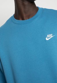 Sudadera Nike azul con un clásico escote redondo y un pequeño logo blanco en el lado izquierdo del pecho. La tela parece suave con una textura lisa.