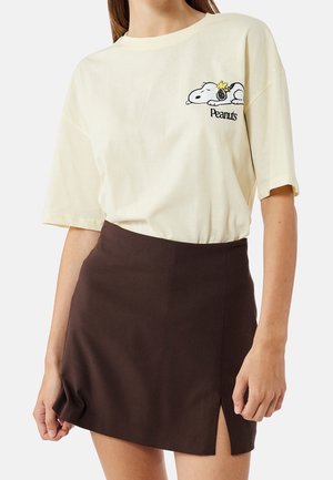Femme portant un t-shirt Peanuts crème avec un motif Snoopy et une mini-jupe noire avec une fente à l'avant.