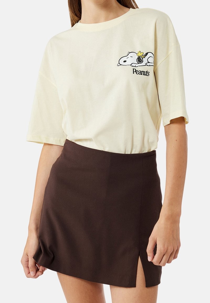 Femme portant un t-shirt Peanuts crème avec un motif Snoopy et une mini-jupe noire avec une fente à l'avant.