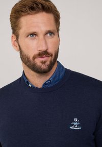 Maglione lavorato a maglia blu navy con scollatura a girocollo, dotato di un ricamo del logo sul lato sinistro del petto e indossato sopra una camicia di denim.