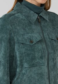 Zielona kurtka z korduroju z zamkiem błyskawicznym, stójką oraz dwoma kieszeniami na piersi z brązowymi guzikami. Widoczna teksturowana tkanina.