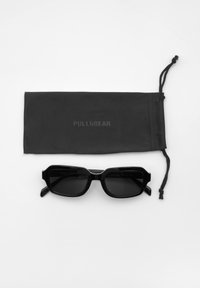 Ochelari de soare rectangulări negri, cu rame groase, însoțiți de un săculeț din țesătură neagră cu un logo subtil.