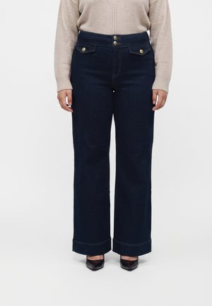 Person iført mørkeblå bukser med vide ben og knapper på forlommerne, beige sweater og sorte spidse sko, stående mod en ensfarvet baggrund.