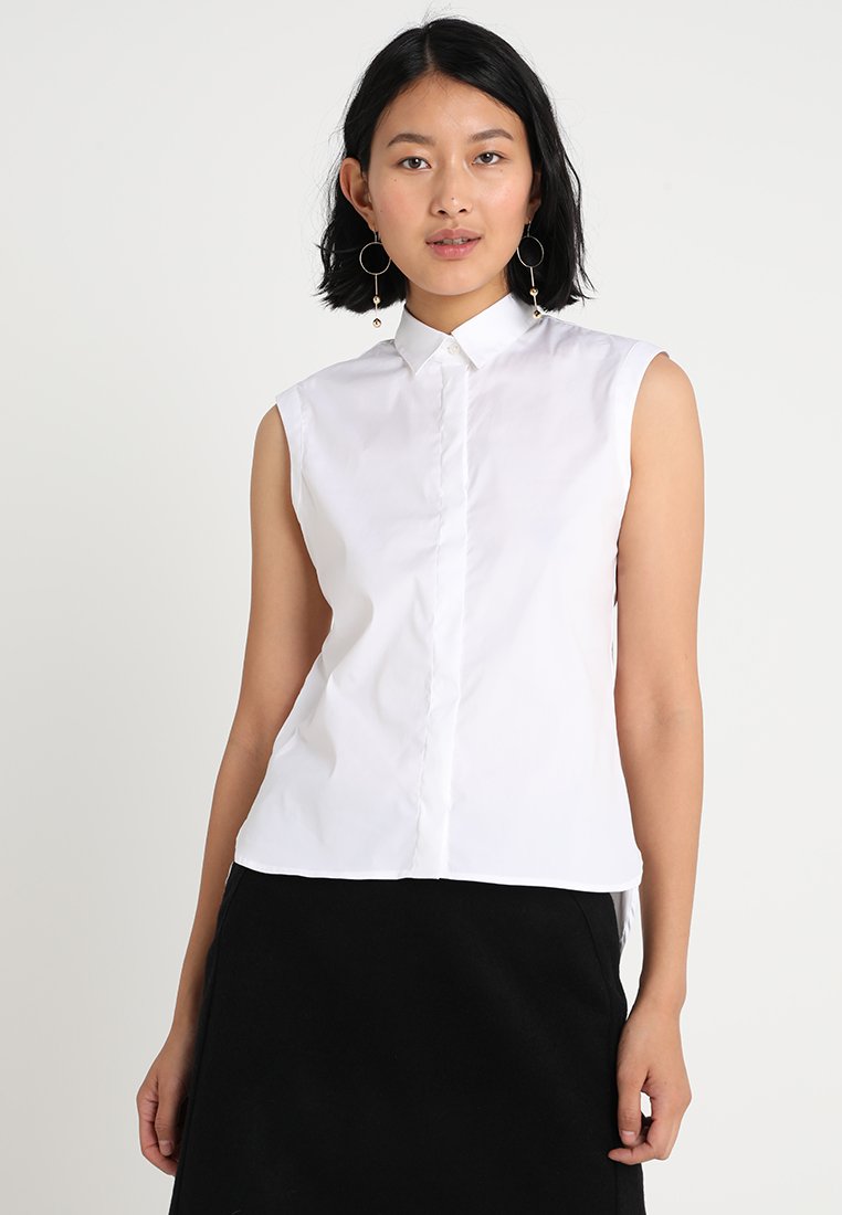 Femme aux cheveux noirs courts portant un chemisier blanc sans manches à col et une jupe noire, debout devant un fond clair uni.