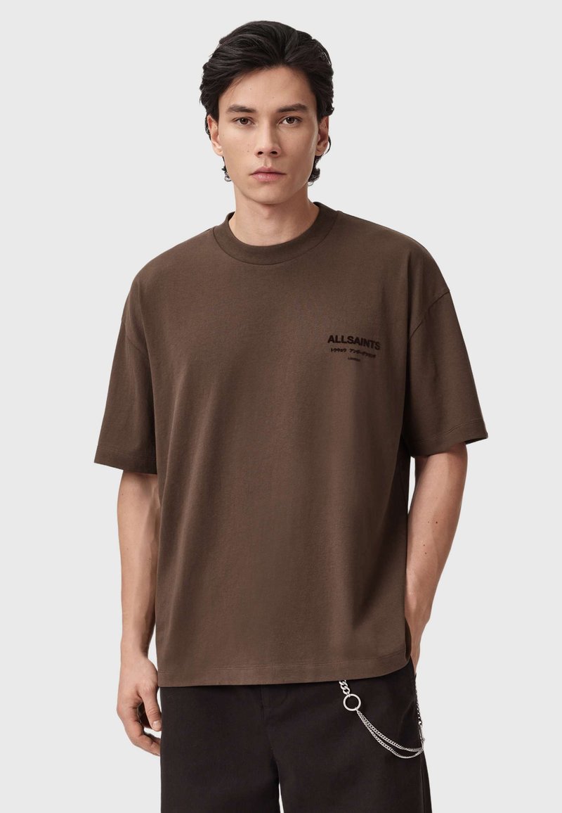Bruine katoenen t-shirt met korte mouwen en een ronde hals. Voorzien van subtiele zwarte "ALLSAINTS" branding op de borst. Gecombineerd met donkere broeken.