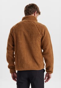 Brun fleecejakke med høj krave, struktureret yderside og en afslappet pasform. Har lange ærmer og et enkelt, sømløst design.