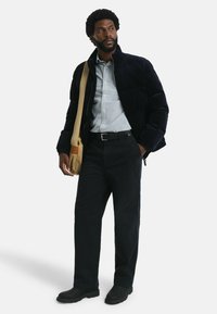 Dunkelblaue Steppjacke mit Kragen, einem karrierten Hemd, schwarze Hose und schwarze Stiefel. Beigefarbenes Schultertasche mit braunen Akzenten.