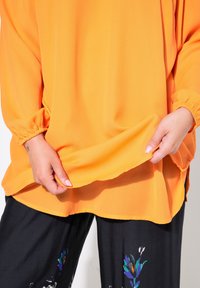 Orangenes Langarmblusenteil mit elastischen Bündchen, glatter Textur und leicht oversized Passform, kombiniert mit schwarzen Hosen mit bunten Blumenmustern.
