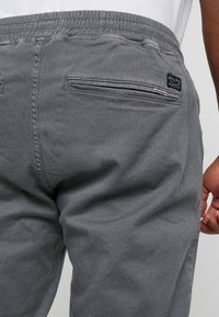 Harmaat housut, joissa on tekstuurillinen kangas, joustava vyötärönauha ja vetoketjullinen takatasku. Pieni merkkilappu on näkyvissä.