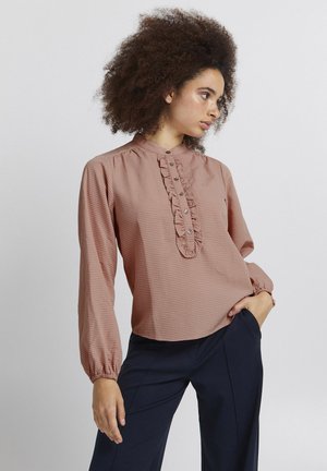 Blouse rose avec un col mandarin, détail volant le long de la patte de boutonnage, et longues manches avec des poignets froncés. Tissu texturé.