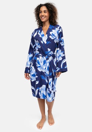 Donna in piedi a piedi nudi che indossa un accappatoio a portafoglio blu navy con grandi motivi floreali bianchi e blu, sorridente e che guarda di lato.