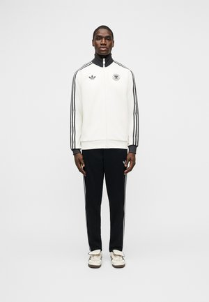 Hombre de pie, vestido con chaqueta blanca con cremallera y rayas negras, pantalones negros con rayas blancas y zapatillas blancas sobre un fondo liso.