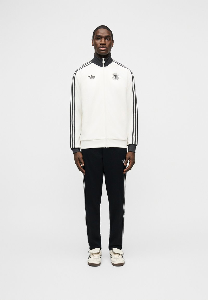 Mand står iført hvid lynlåstrøje med sorte striber og sorte bukser med hvide striber samt hvide sneakers foran en ensfarvet baggrund.