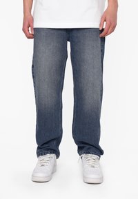 Jeans in denim blu con una finitura leggermente sbiadita, design a gamba dritta e usura sottile. Abbinati a sneakers bianche su uno sfondo neutro.