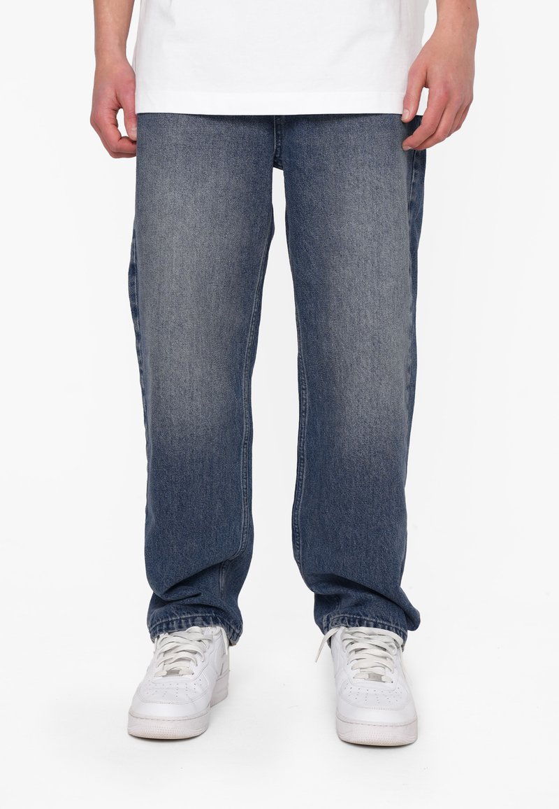 Jeans in denim blu con una finitura leggermente sbiadita, design a gamba dritta e usura sottile. Abbinati a sneakers bianche su uno sfondo neutro.