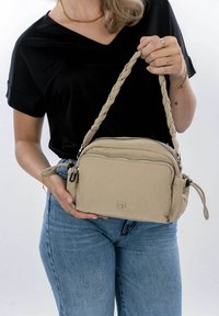 Sac à épaule beige avec une sangle tressée, des compartiments zippés et un logo embossé. Tissu doux avec une texture lisse et une forme arrondie.