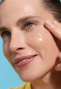 Gel per la cura della pelle applicato sulla guancia di una persona sorridente. Lo sfondo è blu chiaro, mettendo in evidenza i tratti del viso e il prodotto.