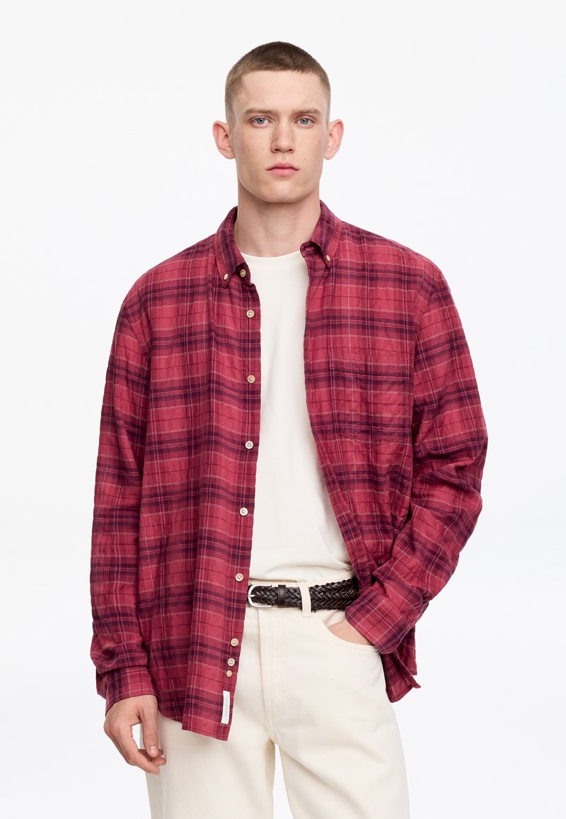 Jeune homme aux cheveux courts portant une chemise à carreaux rouge sur un t-shirt blanc et un pantalon clair avec une ceinture tressée foncée, debout.