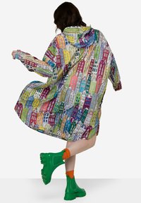 Veelkleurige regenjas met een patroon van geïllustreerde gebouwen, voorzien van een capuchon en een losse pasvorm. Draagt daarbij groene grove laarzen en oranje sokken.