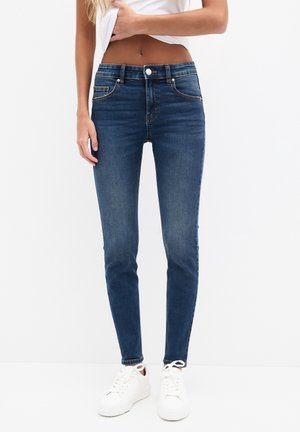 Jean bleu slim porté avec des baskets blanches et un top blanc court, montrant le ventre et une posture décontractée sur fond uni.