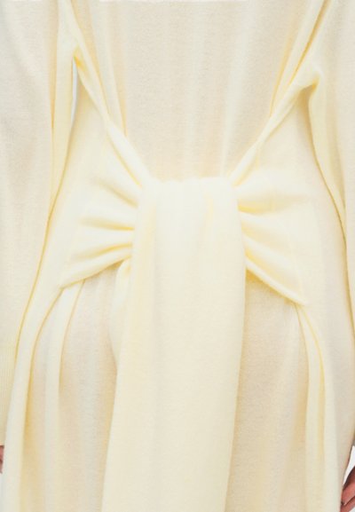 Απαλό κίτρινο πλεκτό ένδυμα με φαρδιά, συγκεντρωμένη ζώνη δεμένη κόμπο στη μέση, που αναδεικνύει την υφή και την πτώση του υφάσματος.