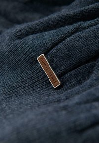 Pull en tricot bleu foncé avec un tissu texturé. Présente une petite étiquette en métal portant l'inscription "QUINTESSENCE" attachée sur le côté.