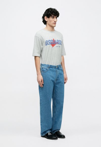 Hall halli lühikeste varrukatega T-särk sinise ja punase graafilise tekstiga, kombineeritud lahtiste heleroheliste teksade ja mustade kingadega, seisab lihtsal taustal.