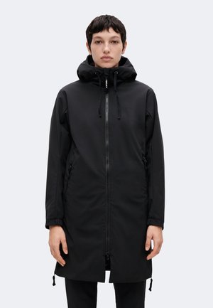 Personne portant une longue veste noire à capuche avec fermeture éclair sur le devant et des poches latérales zippées, debout devant un fond clair uni.