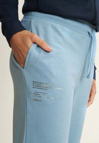 Helle blaue Sweatpants mit einer weichen Textur, ausgestattet mit einer Seitentasche und einem goldenen Folientextdetail am linken Oberschenkel. Mit Kordelzugbund.